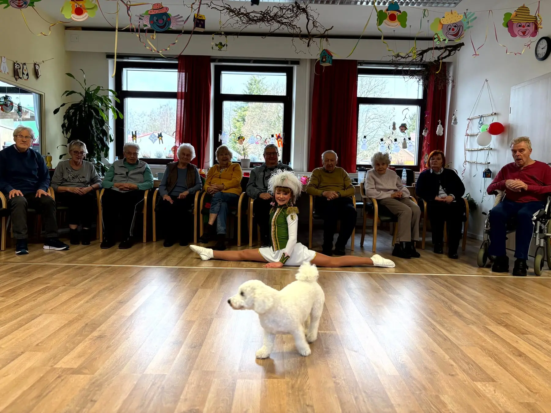 Gäste sitzen im großen Aufenthaltsraum der Tagespflege Weißdorf im Stuhlkreis, Tagespflegehund Luna läuft durch die Mitte